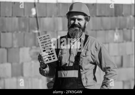 Elemento in muratura. Builder tenere mattone. Uomo con bearded indossa un elmetto in cantiere. Materiale di costruzione di mattone usato per fare le pareti Foto Stock