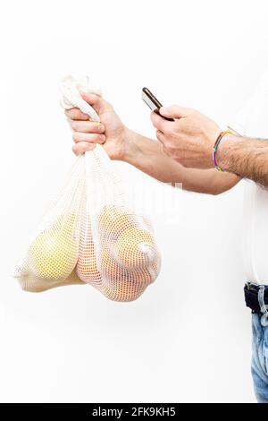 Un uomo irriconoscibile che tiene alcuni sacchetti di rete organici con le mele e le arance mentre guarda il suo telefono delle cellule. Foto Stock