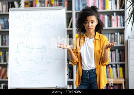 Ritratto di giovane donna afro-americana confusa in piedi vicino a lavagna bianca, in abiti casual, conduce lezioni, comunicazione online con gli studenti, guardando la macchina fotografica a sorpresa Foto Stock