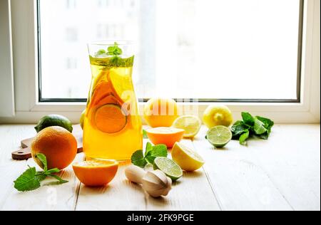 Caraffa di limonata ghiacciata, centrifuga in legno, acqua fredda infusa di agrumi con fette di limone e lime, foglie di menta, tagliere su davanzale in legno bianco. A parte Foto Stock