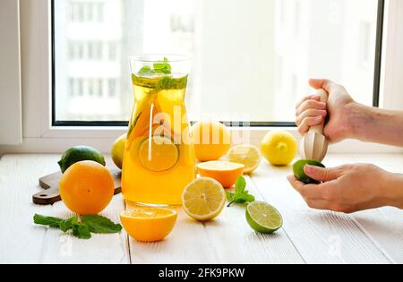 Primo piano delle mani di una giovane donna che fanno limonata fresca, spremono il succo dagli agrumi, spremono. Caraffa piena di bevanda fredda con limone, arancia, l Foto Stock