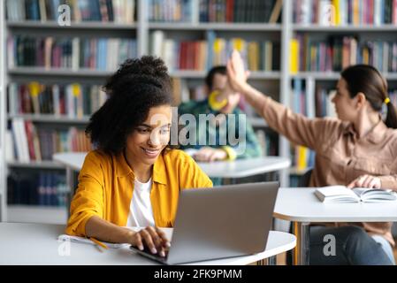 Felice sicuro ragazza teenage afro-americana intelligente seduta al tavolo in biblioteca, utilizzando il computer portatile mentre si prepara per le lezioni o l'esame, due studenti sfocati si danno cinque in background Foto Stock