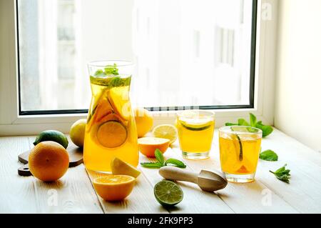 Caraffa di limonata ghiacciata e due bicchieri, centrifuga in legno, acqua fredda infusa di agrumi, limone e lime, foglie di menta, tagliere su davanzale in legno bianco. AP Foto Stock