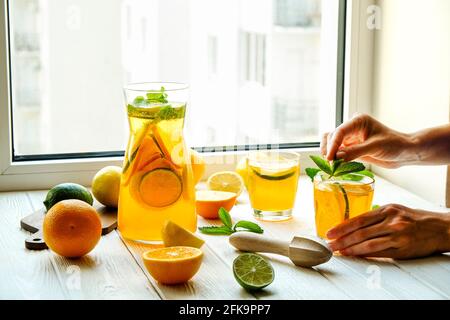 Primo piano delle mani di una giovane donna che fanno limonata fresca, spremono il succo dagli agrumi, spremono. Caraffa piena di bevanda fredda con limone, arancia, l Foto Stock