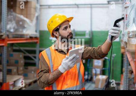 Uomo giovane e serio in hardhat e guanti scansione codice dell'articolo mentre fa l'inventario di stock al deposito Foto Stock