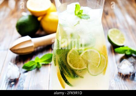 Caraffa di limonata ghiacciata, centrifuga in legno, bevanda fredda all'acqua infusa di agrumi con fette di limone e lime, foglie di menta su tavola testurizzata di legno brunito marrone su Foto Stock