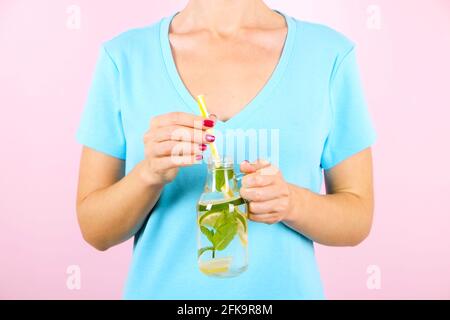 Primo piano di mani di giovane donna che tengono la limonata appena spremuta di agrumi. Caraffa piena di bevande fredde con foglie di limone, arancia, lime e menta Foto Stock