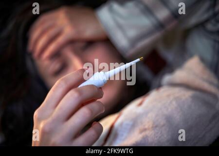 Giovane donna offuscata che si sente malata, avendo febbre guardando il termometro che tocca la fronte. Concetto di malattia e febbre. Foto Stock