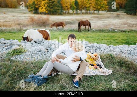 La coppia è seduta abbracciandosi su una coperta con cibo sul prato. I cavalli pascolano sullo sfondo Foto Stock