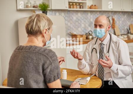 Un medico di sesso maschile che indossa una maschera protettiva consiglia una donna anziana che indossa una maschera protettiva a casa sua. Foto Stock