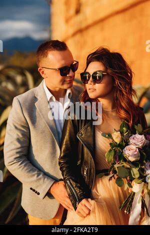 Lo sposo si trova dietro la sposa in occhiali da sole e una giacca in pelle con un bouquet di fiori sullo sfondo del muro del castello Foto Stock