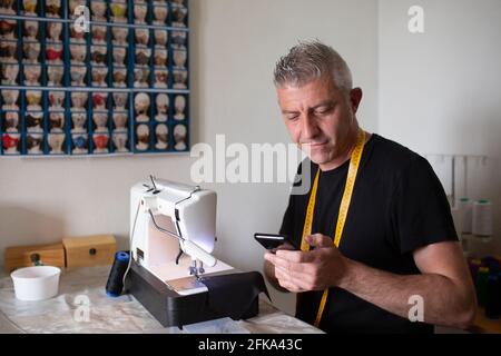 un sarto che lavora con la sua macchina da cucire consultando il suo mobile telefono Foto Stock