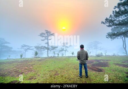 L'uomo viaggiatore si erge a guardare l'alba su una nebbiosa collina di pini del mattino sulla cima delle alture di da Lat, in Vietnam Foto Stock