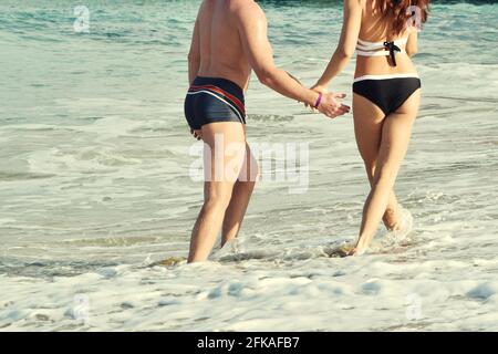 In spiaggia una giovane coppia che corre in mare ondeggia in costume da bagno. Braccialetto dell'hotel tutto compreso a portata di mano Foto Stock