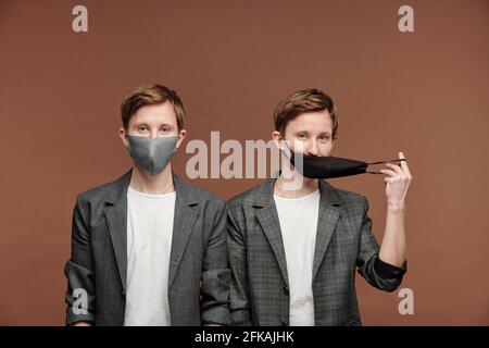 Ritratto di bei fratelli gemelli in eleganti abiti grigi che si posano in maschere su sfondo marrone, uno di loro tirando maschera Foto Stock