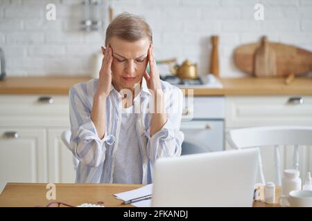Sensazione di malessere. Donna matura stressata con gli occhi chiusi che soffrono di mal di testa mentre si siede in cucina a casa, laptop e farmaci su tavolo di legno. Concetto di assistenza sanitaria e persone anziane Foto Stock