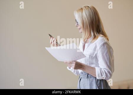 Fai in modo che il business accada. Donna matura occupata con capelli biondi in abbigliamento di affari che tiene il suo smartphone e carte, in piedi all'interno Foto Stock