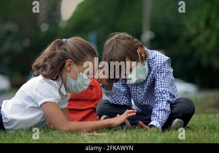 Studenti carini che usano gli smartphone alla scuola elementare. Ragazzo e ragazza in maschere di sicurezza con gadget nelle loro mani. Foto Stock