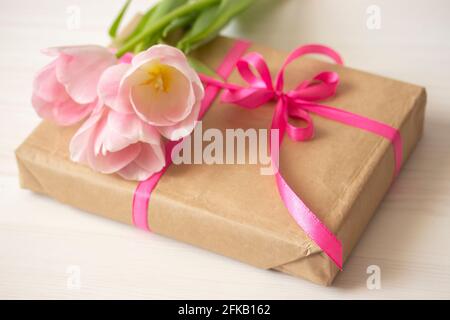 Regalo avvolto in carta artigianale e legato con un nastro rosa e un bouquet di delicati tulipani rosa. Piccolo regalo da vicino Foto Stock