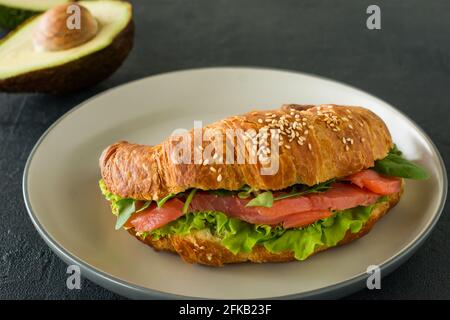 Panino di croissant con salmone salato su un piatto, servito con foglie fresche di insalata, rucola e verdure su sfondo nero. Foto Stock