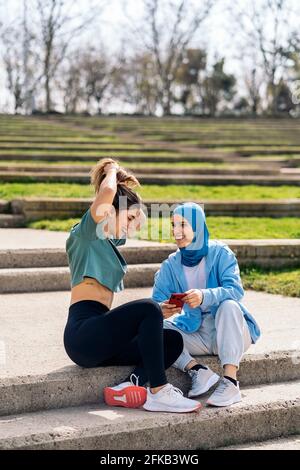 Felice ragazza musulmana e la sua amica seduta nel parco che parla e usa il telefono cellulare. Foto Stock