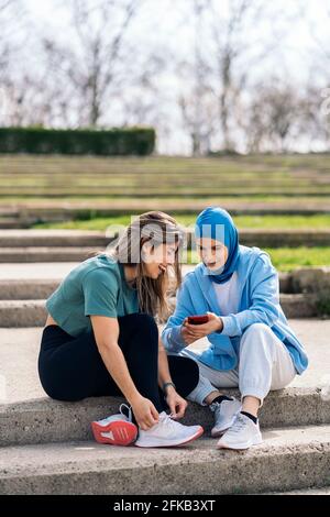 Felice ragazza musulmana e la sua amica seduta nel parco che parla e usa il telefono cellulare. Foto Stock