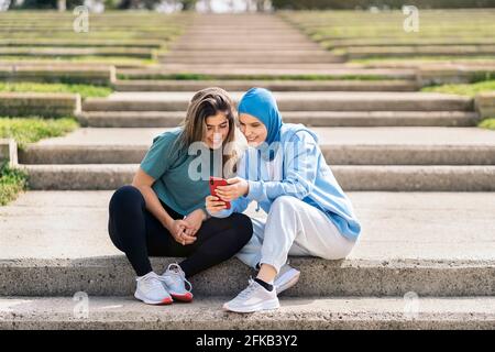 Felice ragazza musulmana e la sua amica seduta nel parco che parla e usa il telefono cellulare. Foto Stock