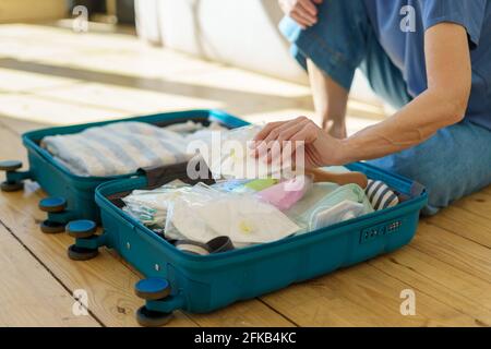 Donna non riconoscibile tagliata che si prepara a viaggiare durante l'epidemia di coronavirus, mettendo respiratore o maschera facciale in valigia prima del viaggio di vacanza in tempo pandemico. Concetto di viaggio sicuro Foto Stock