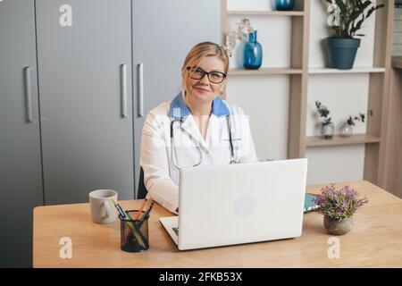 Buon dottore biondo senior sorridente alla macchina fotografica mentre si usa un computer portatile in ufficio Foto Stock