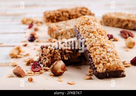 Varie barrette energetiche di cioccolato alla granola in fila con noci miste sparse, cereali e frutta secca, sfondo di tavola di legno bianco grunged. Sano vegan fitn Foto Stock