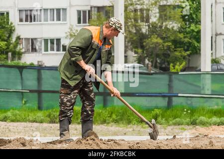 Bielorussia, Minsk - 28 maggio 2020: Operaio industriale maschile scavando terra con pala e livellamento con rastrello in cantiere. Foto Stock