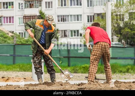 Bielorussia, Minsk - 28 maggio 2020: Lavoratori industriali maschi scavare terra con pala e livellamento con rastrello in cantiere. Foto Stock