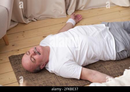 Uomo anziano meditating su un pavimento di legno del soggiorno e sdraiato in Shavasana Foto Stock