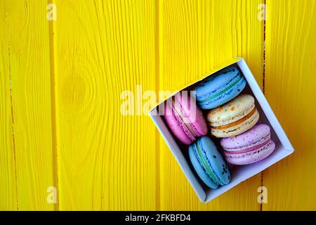 Dolci alla amarina francese, gusti e colori diversi. Scatola di macaron su sfondo tavola di legno giallo tessitura legno. Compleanno San Valentino regalo di primavera Foto Stock