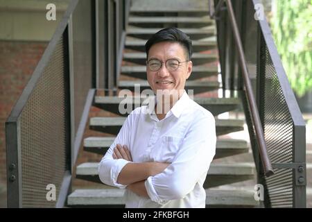 Ritratto di un uomo d'affari asiatico sorridente con le braccia incrociate, con camicia bianca. Scala sfondo. Foto Stock