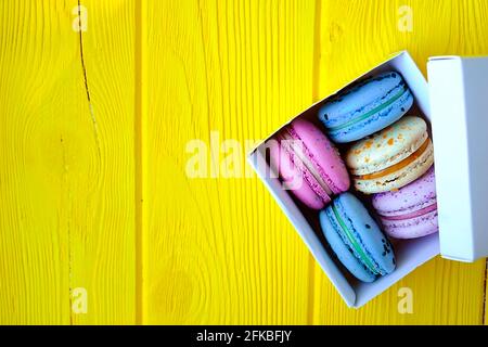 Dolci alla amarina francese, gusti e colori diversi. Scatola di macaron su sfondo tavola di legno giallo tessitura legno. Compleanno San Valentino regalo di primavera Foto Stock