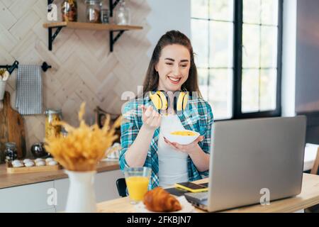 Una giovane donna che fa colazione comunica con una videochiamata in un computer portatile in cucina. La ragazza lavora da casa, consultazioni online e lezioni. Tu Foto Stock