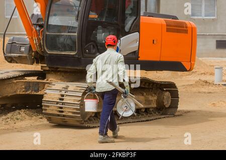 Il lavoratore industriale in un casco rosso cammina con un utensile di lavoro sullo sfondo di un escavatore cingolato in un cantiere. Foto Stock