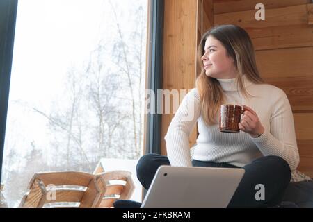 Donna che indossa una camicia a collo alto seduta a gambe incrociate che tiene una tazza da caffè vicino a un computer portatile in una stanza accogliente. Guardando attraverso la finestra e sorridendo. Giornata rilassante a casa tua. Foto Stock