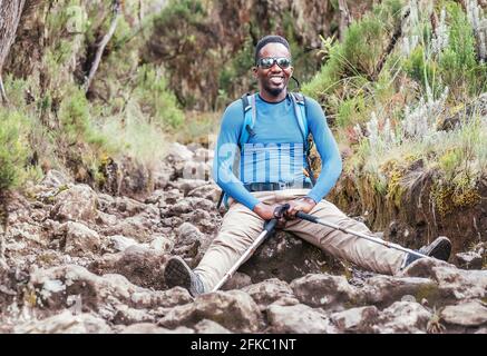 Ritratto di un allegro sorriso afroamericano Ethnicity giovane in occhiali da sole seduti con zaino e bastoni da trekking e su cui riposarsi montagna t Foto Stock