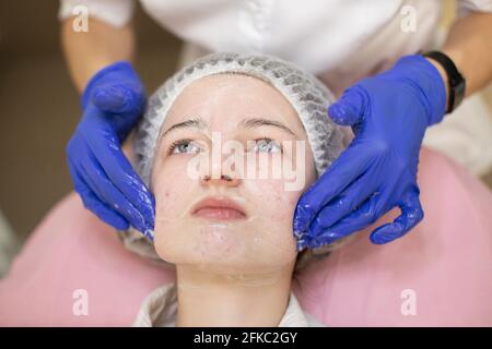 Primo piano di mani di cosmetologo in guanti di gomma blu, applicazione professionale peeling o schiuma detergente per la giovane ragazza viso. Trattamento dell'acne e del pimple della pelle alla clinica moderna Foto Stock