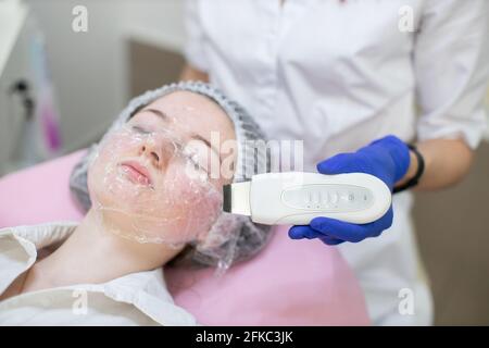 Le mani di donna beautician medico con un dispositivo per la pulizia ad ultrasuoni vicino al volto di teen ragazza in cappello monouso, con pellicola sul viso. Trattamento dell'acne, concetto di pelle problematico Foto Stock