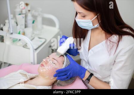 Concetto di cosmetologia e cura professionale della pelle, trattamento dell'acne. Primo piano di cosmetologist femminile che fa procedura di pulizia ultrasonica faccia per la ragazza teen con pelle di acne in un salone di bellezza Foto Stock