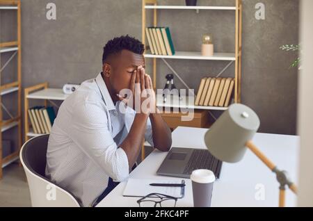 Uomo nero seduto alla scrivania del suo ufficio sentendosi stanco, sopraffatto e stressato fuori Foto Stock