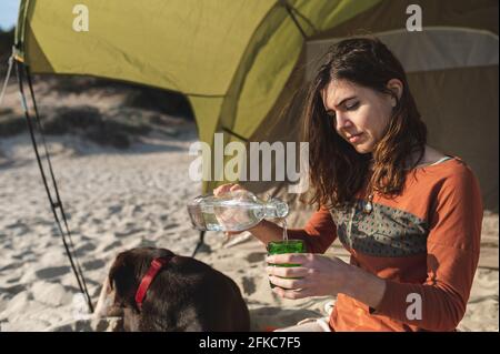 Primo piano giovane donna attraente in abbigliamento casual versando acqua in vetro. Cane che dorme sullo sfondo. Stile di vita in campeggio. Foto Stock