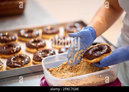 Un lavoratore spruzza nocciole macinate su ciambelle in un'atmosfera di lavoro in un laboratorio di caramelle. Pasticceria, dessert, dolce, fare Foto Stock