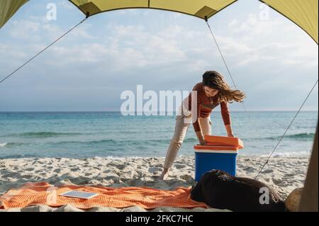 Vista dall'interno della tenda di giovane donna in un campeggio sulla spiaggia che apre il frigorifero. Giornata ventosa e soleggiata. Stile di vita all'aperto. Foto Stock