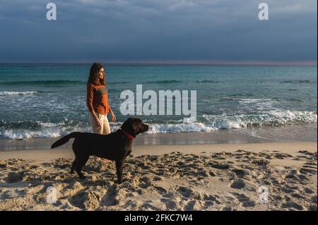 Donna in piedi sulla spiaggia guardando l'orizzonte con il suo cane che guarda la stessa direzione Foto Stock