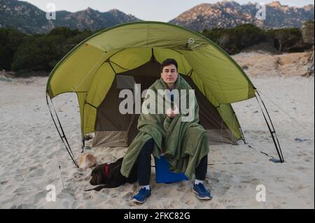 Giovane uomo che indossa una coperta per riscaldarsi durante una tenda da campeggio sulla spiaggia con il suo cane. Foto Stock