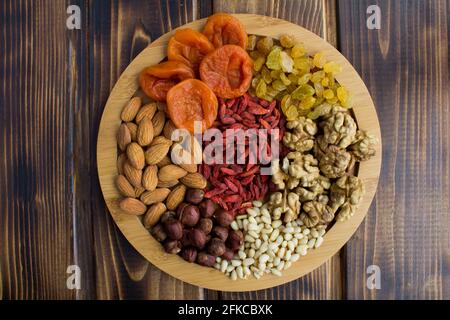 Albicocche secche, uvetta, bacche di goji, noci diverse sul tagliere rotondo sul legno marrone background.Top vista. Foto Stock
